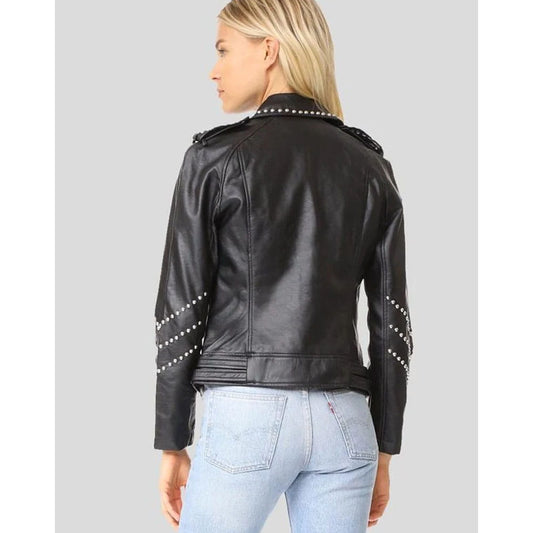 A woman shown from the back wearing a black faux-leather moto jacket with silver stud detailing on the shoulders and sleeves, paired with light-wash blue jeans, against a solid gray background.