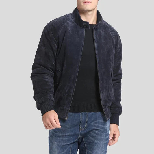 A man wearing a dark navy blue suede or velvety bomber jacket over a black sweater and blue jeans, photographed against a solid light gray background.