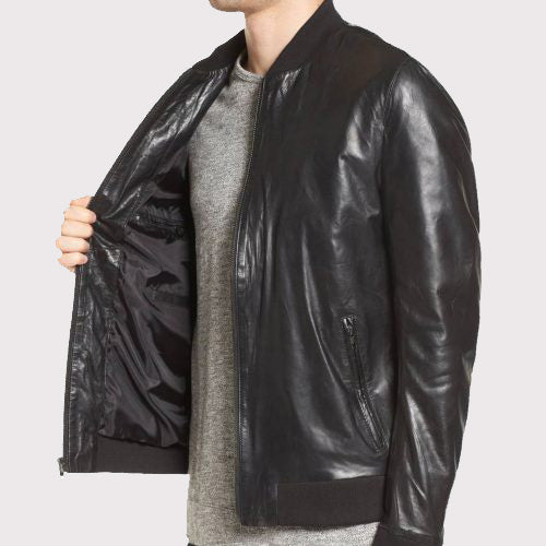 A man wearing a black leather bomber jacket over a gray crewneck sweater, shown from the neck down against a light gray background. He is holding the jacket open to show the black interior lining.