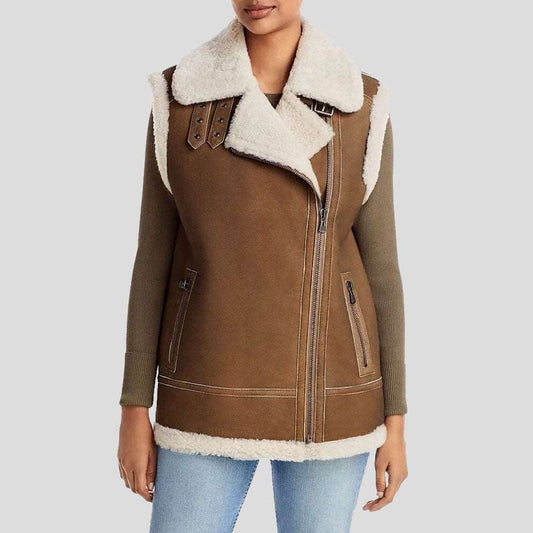 A woman wearing a brown shearling-lined aviator-style vest with olive green knit sleeves and light blue jeans, shown against a plain light gray studio background.