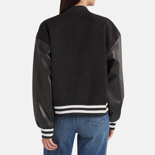 A person shown from the back wearing a black varsity-style jacket with black faux-leather sleeves and white striped ribbing, paired with blue jeans against a light gray studio background.