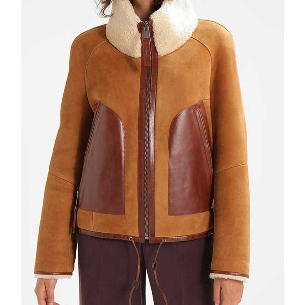 A cropped view of a person wearing a tan and brown shearling-style jacket with a cream sherpa collar, front zipper, and dark brown pants against a light gray studio background.