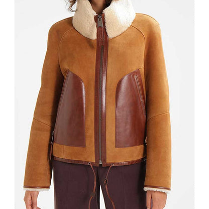 A cropped view of a person wearing a tan and brown shearling-style jacket with a cream sherpa collar, front zipper, and dark brown pants against a light gray studio background.