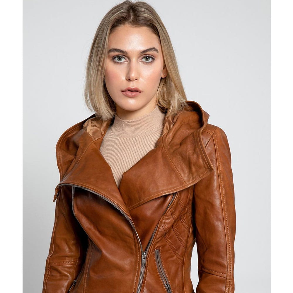 A woman wearing a brown leather moto jacket with an asymmetrical zipper and a high collar, paired with a beige ribbed turtleneck sweater, set against a plain light gray background.