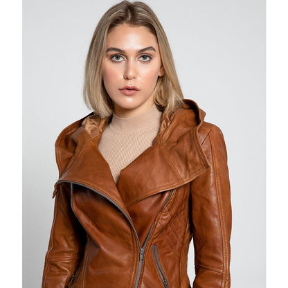 A woman wearing a brown leather moto jacket with an asymmetrical zipper and a high collar, paired with a beige ribbed turtleneck sweater, set against a plain light gray background.