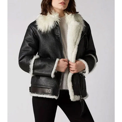 A woman wearing a black faux-leather aviator-style jacket with a thick white shearling-style collar, cuffs, and hem, paired with a light gray sweater and black pants, against a plain light gray studio background.