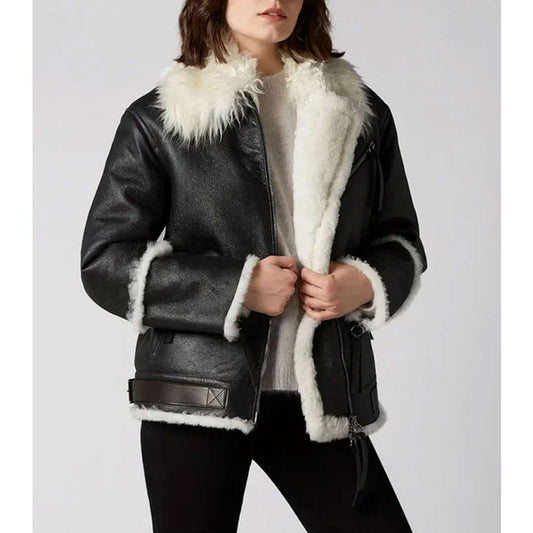 A woman wearing a black faux-leather aviator-style jacket with a thick white shearling-style collar, cuffs, and hem, paired with a light gray sweater and black pants, against a plain light gray studio background.