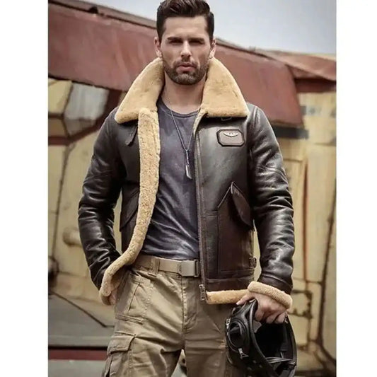 A man wearing a dark brown leather aviator jacket with a beige shearling lining, holding a black motorcycle helmet. He is wearing tan cargo pants and a dark gray t-shirt, standing in front of a rustic industrial building.