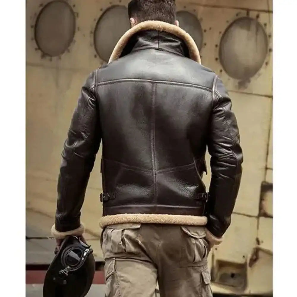 A man viewed from behind wearing a dark brown leather aviator jacket with a beige shearling lining, paired with khaki cargo pants and holding a black helmet, set against a vintage aircraft fuselage background.
