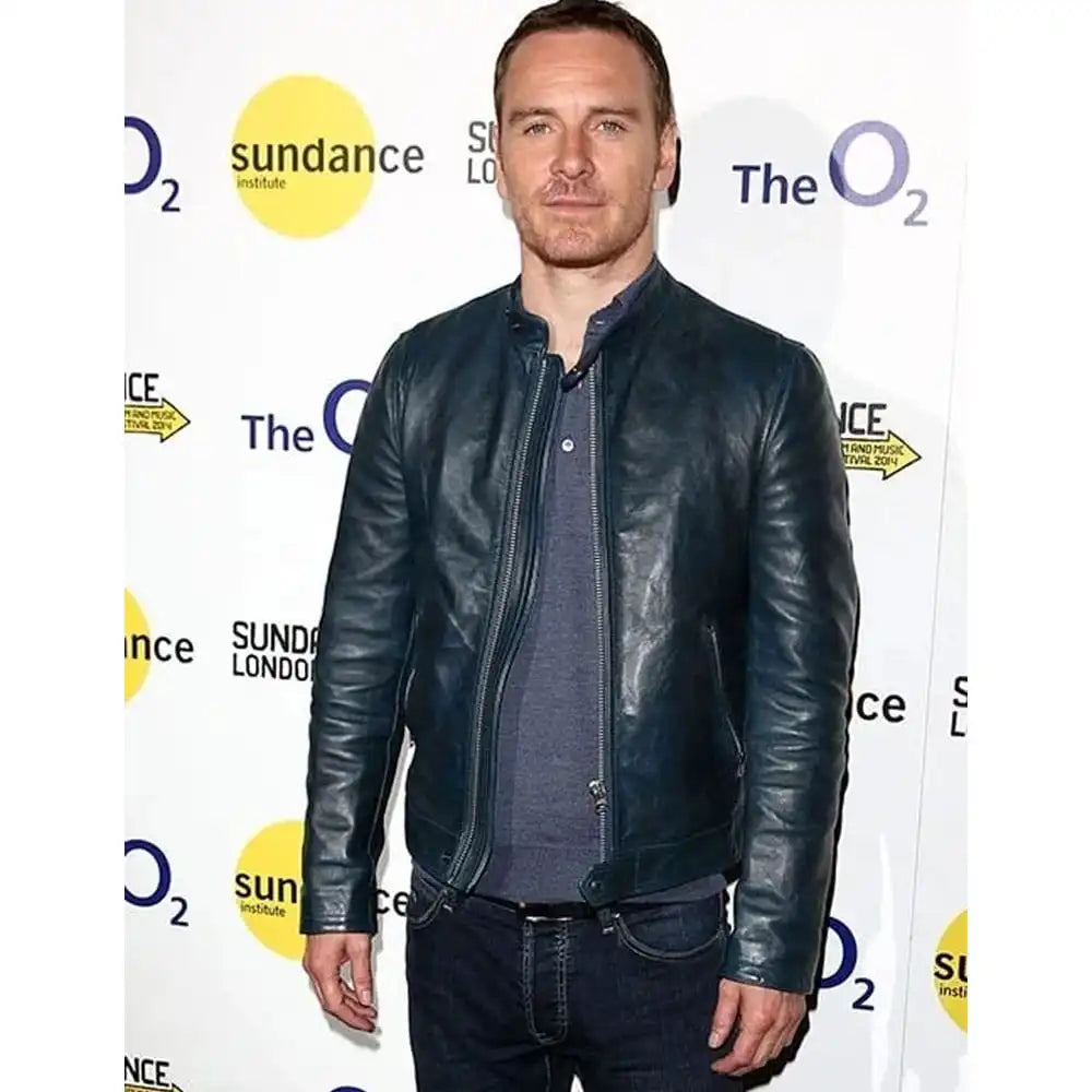 A man wearing a dark navy blue leather jacket over a dark gray shirt and jeans, standing in front of a white event backdrop with 'sundance institute' and 'The O2' logos.