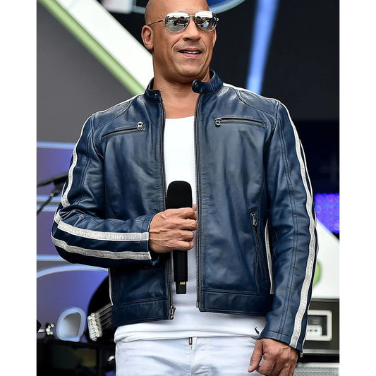 A man wearing a dark blue leather jacket with white stripes on the sleeves, a white t-shirt, and white pants, holding a microphone on stage.