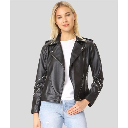 A woman wearing a black faux leather moto jacket with silver studs on the shoulders and sleeves, styled over a white t-shirt and light blue jeans, set against a solid gray background.