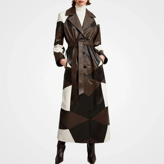 A model wears a long geometric patchwork trench coat made of brown, black, and off-white panels, styled with a white turtleneck and black boots against a light gray studio background.