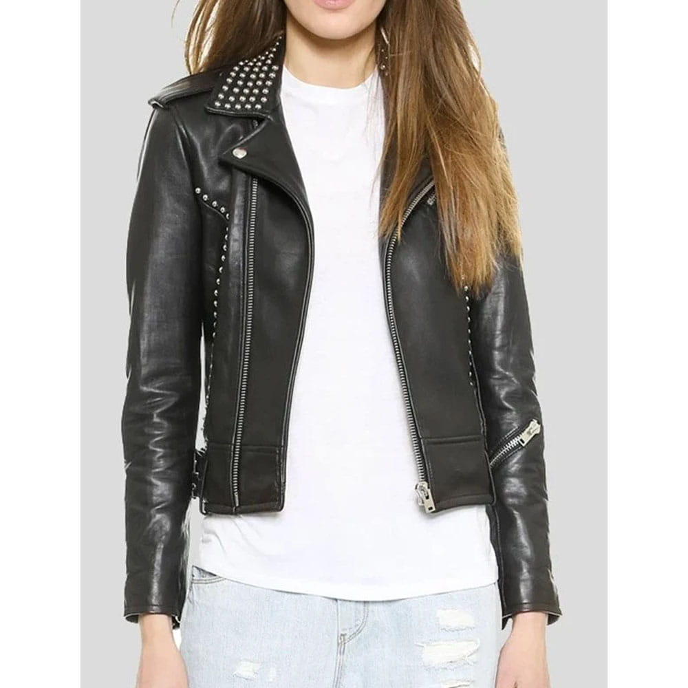 A woman wearing a black faux-leather moto jacket with silver studs and zippers, layered over a white t-shirt and light-wash distressed jeans, shown against a solid gray background.