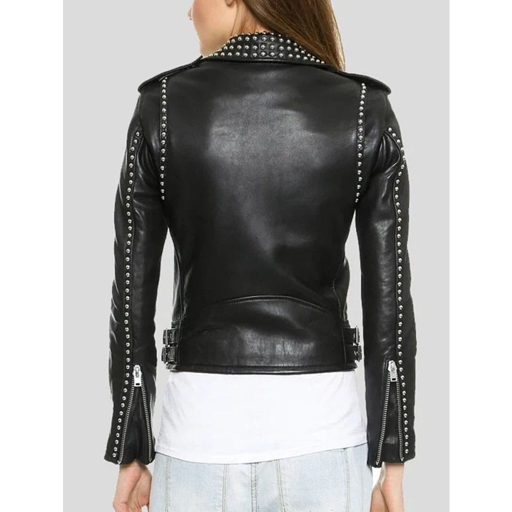 A woman shown from the back wearing a black faux-leather moto jacket with silver studs and zippers, layered over a white t-shirt and light blue jeans, against a solid gray background.