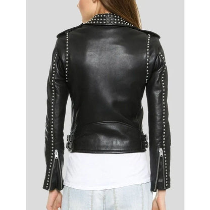 A woman shown from the back wearing a black faux-leather moto jacket with silver studs and zippers, layered over a white t-shirt and light blue jeans, against a solid gray background.