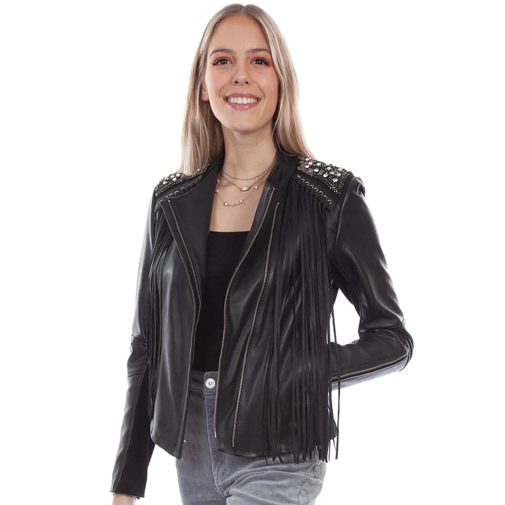 A woman modeling a black faux-leather moto jacket with fringe detailing and studded shoulder accents, paired with a black top and gray jeans against a white background.