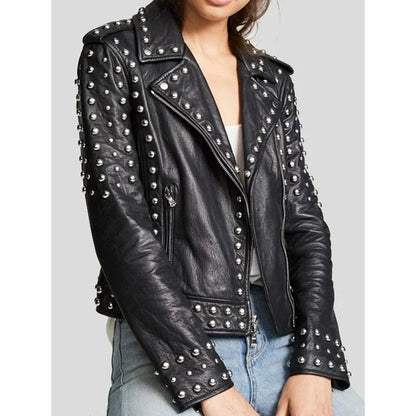 A woman wearing a black leather moto jacket heavily decorated with silver studs, paired with light blue jeans against a solid gray background.
