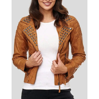 A woman wearing a cropped tan faux-leather moto jacket with silver studs and zippers, layered over a white t-shirt and black pants, against a solid gray background.