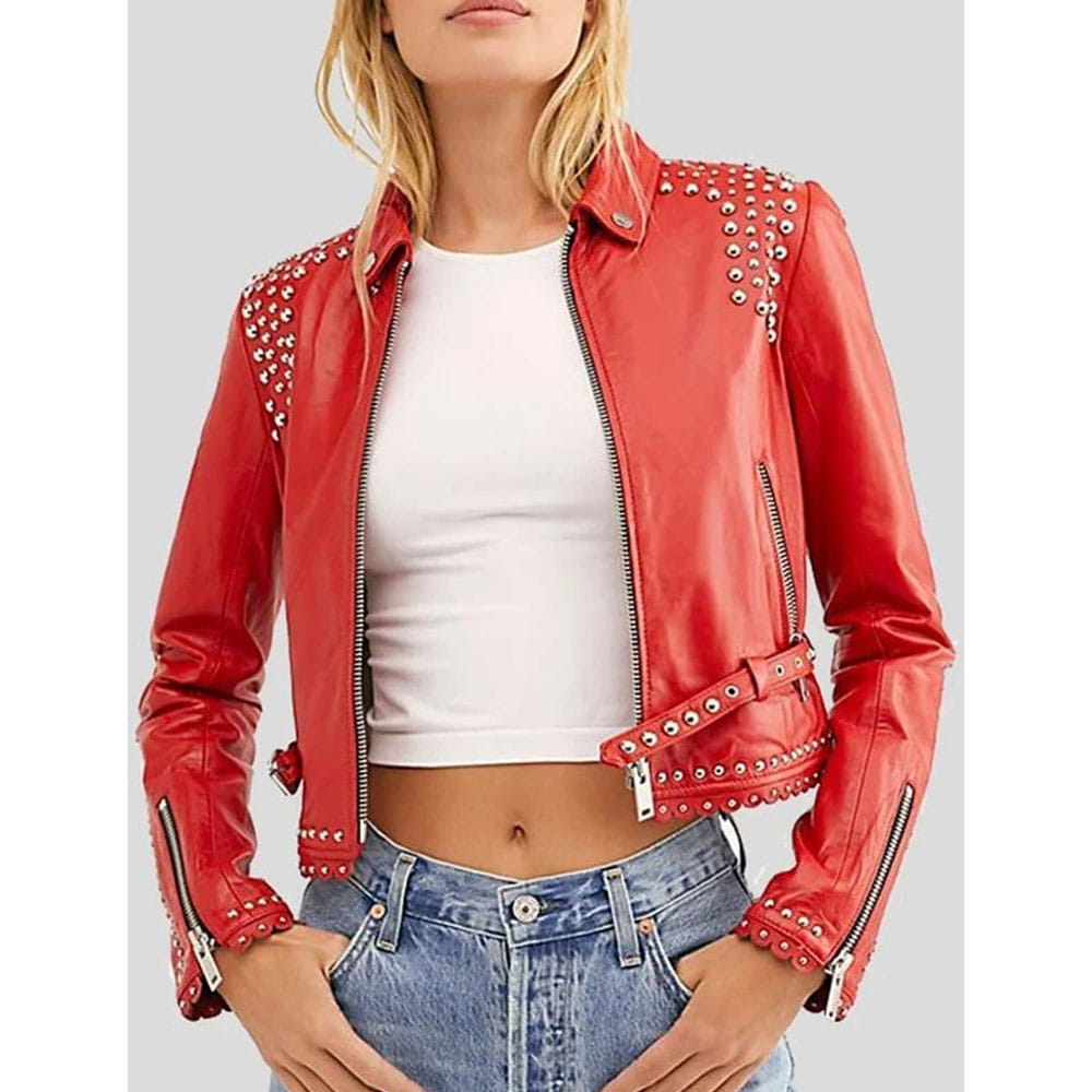 A woman wearing a bright red faux leather moto jacket with silver studs on the shoulders and cuffs, styled with a white crop top and blue jeans against a solid gray background.