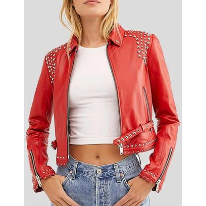 A woman wearing a bright red faux leather moto jacket with silver studs on the shoulders and cuffs, styled with a white crop top and blue jeans against a solid gray background.