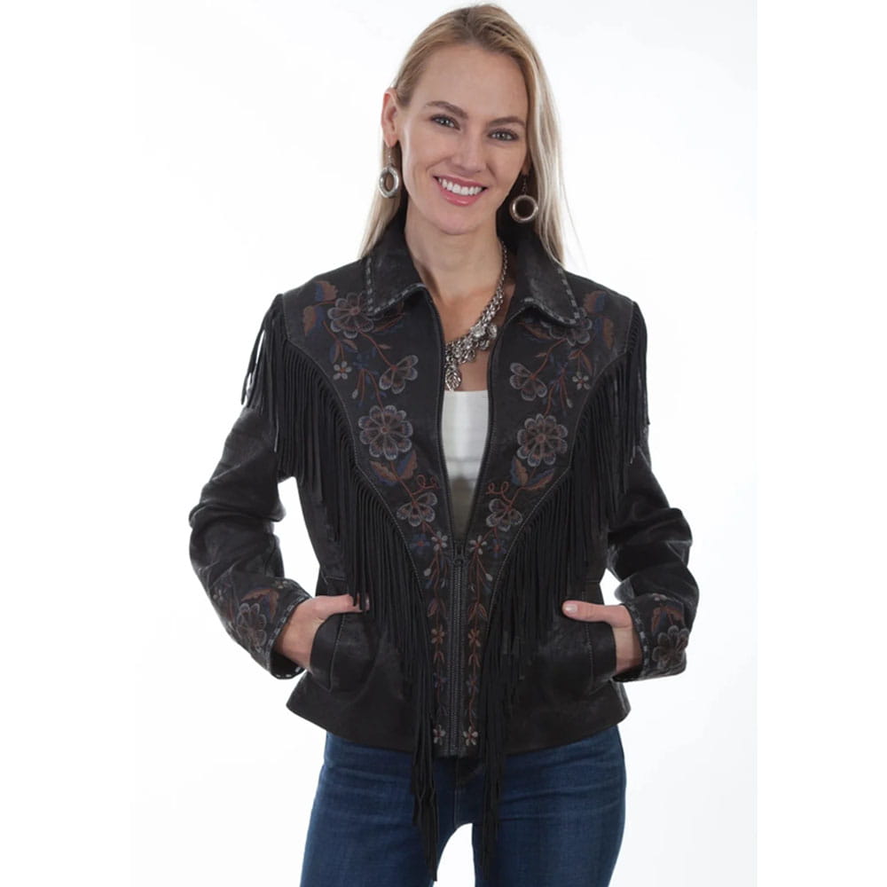 A woman modeling a black suede fringe jacket with intricate floral embroidery on the front and sleeves, styled with blue jeans and a statement necklace against a plain white background.