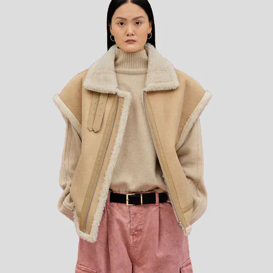 A model wearing a beige shearling-lined jacket over a beige turtleneck sweater, paired with pink pants and a black belt, against a solid light gray background.