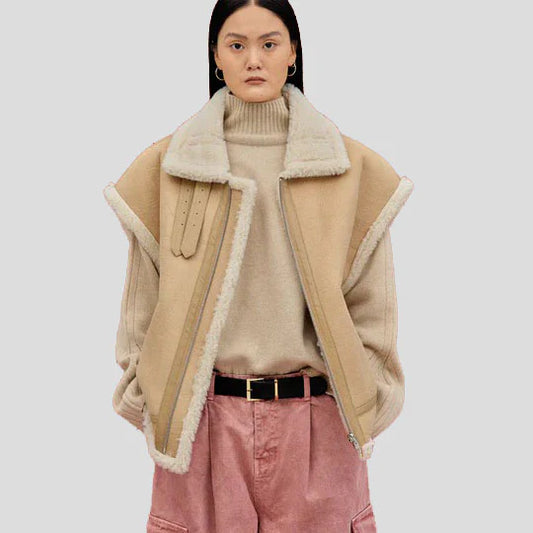 A model wearing a beige shearling-lined jacket over a beige turtleneck sweater, paired with pink pants and a black belt, against a solid light gray background.