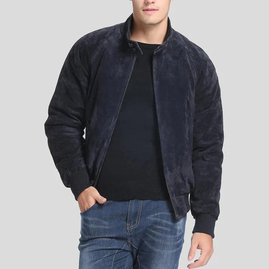 A man wearing a dark navy blue suede or velvety zip-up jacket over a black sweater and blue jeans, shown from the chin down against a plain light gray background.