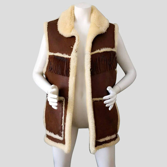 A brown and beige shearling-style sleeveless vest displayed on a white mannequin against a light gray background. The vest features dark brown leather-like panels, cream-colored shearling trim, fringe details, and large front pockets.
