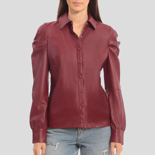 A woman wearing a fitted long-sleeve button-up blouse in a deep burgundy or maroon faux leather, paired with light-wash distressed jeans, against a solid light gray background.