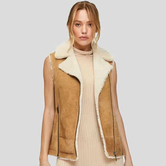 A woman wearing a tan suede shearling-lined sleeveless vest over a cream ribbed turtleneck sweater, photographed against a solid light gray studio background.