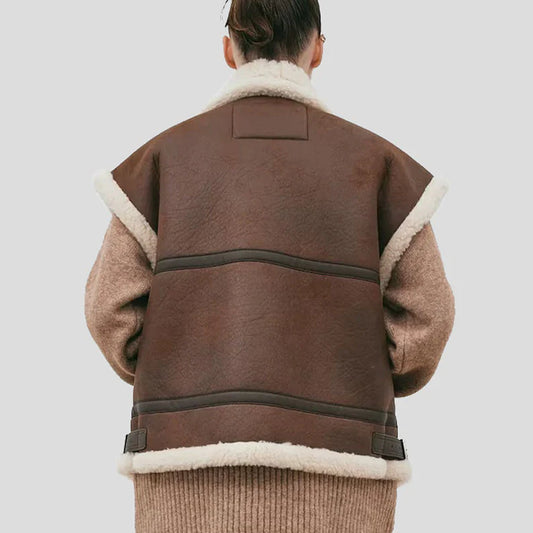 Rear view of a person wearing a brown shearling-lined jacket with beige knit sleeves and a cream-colored fleece collar, set against a solid light gray background.