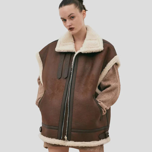 A model wearing a brown faux-shearling jacket with cream-colored sherpa lining on the collar, cuffs, and pockets, set against a solid light gray background.