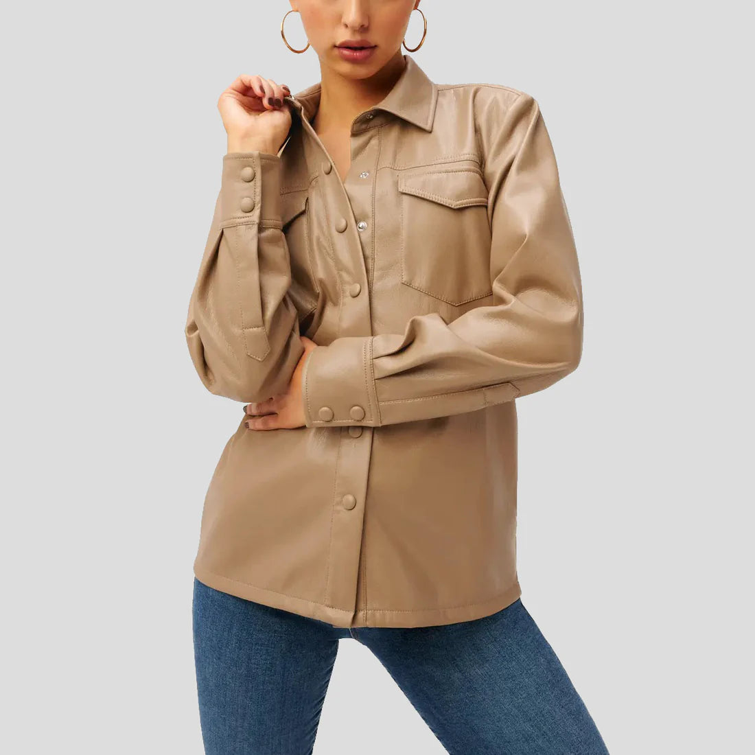 A woman wearing a beige long-sleeve button-up shirt with chest flap pockets and blue jeans, shown from the mouth down against a light gray studio background.
