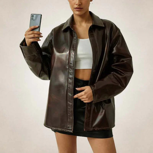 A woman takes a mirror selfie wearing an oversized dark brown faux-leather jacket over a white crop top and black shorts, holding a smartphone.
